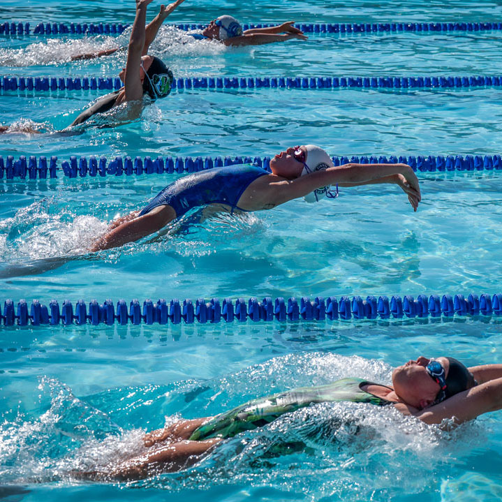 Young swimmers launch from the starting blocks backwards in a backstroke race.
